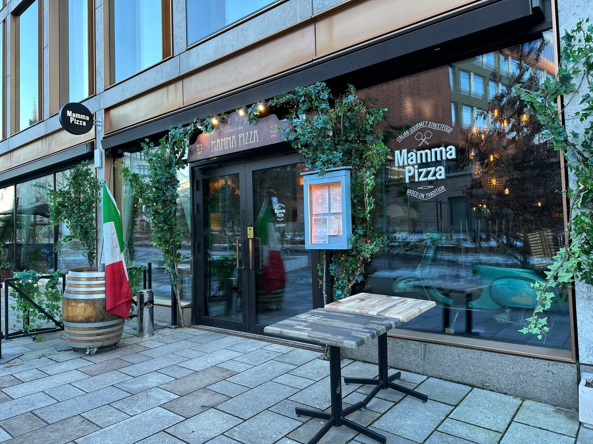 Mamma Pizza's greenery-framed entrance with Italian flags and barrel table