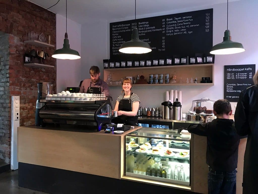 Baristas behind the counter at Supreme Roastworks coffee bar in Grünerløkka, Oslo, with espresso machine and pastry case.